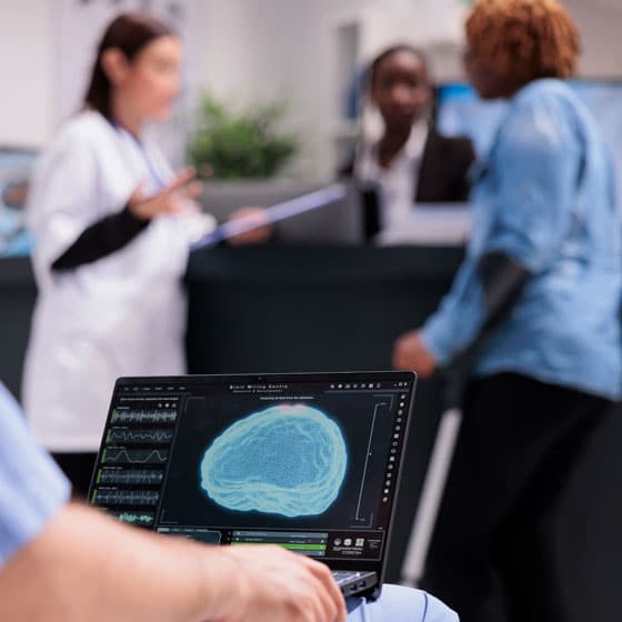 Healthcare professional reviewing a brain scan on a laptop in a clinical setting while medical staff discuss patient care in the background.